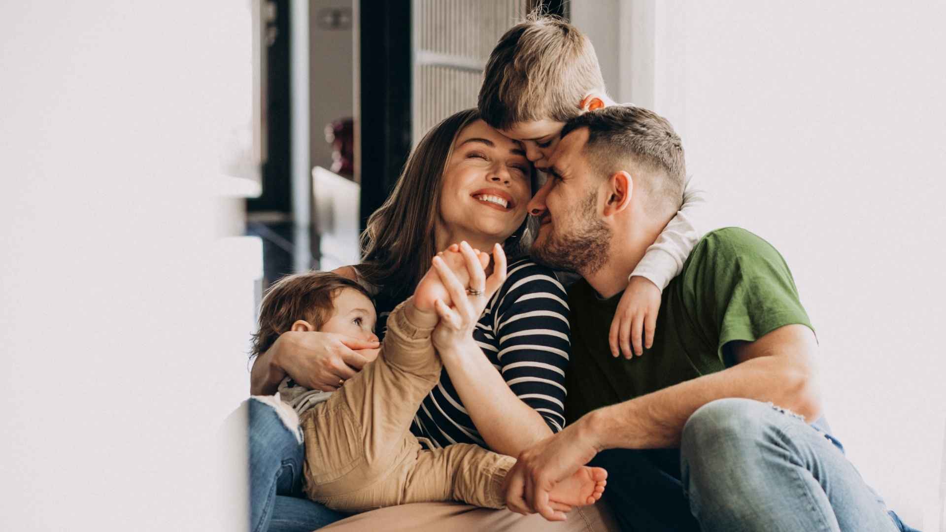 Eine glückliche vierköpfige Familie sitzt eng beieinander in einem Haus, lächelt und umarmt sich. Die Mutter und der Vater halten sich an den Händen, während ihre beiden kleinen Kinder sich warm an sie schmiegen. Das Sonnenlicht fällt von der linken Seite ein.
