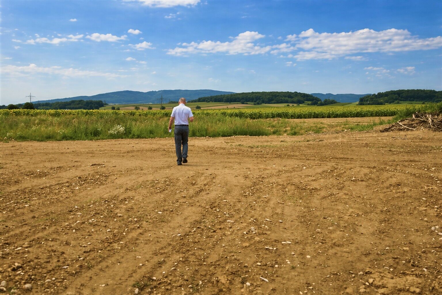 Ein älterer Mann geht allein auf einem trockenen, unbefestigten Feld in Richtung grüner Vegetation und entfernter Hügel unter einem blauen Himmel mit vereinzelten Wolken.