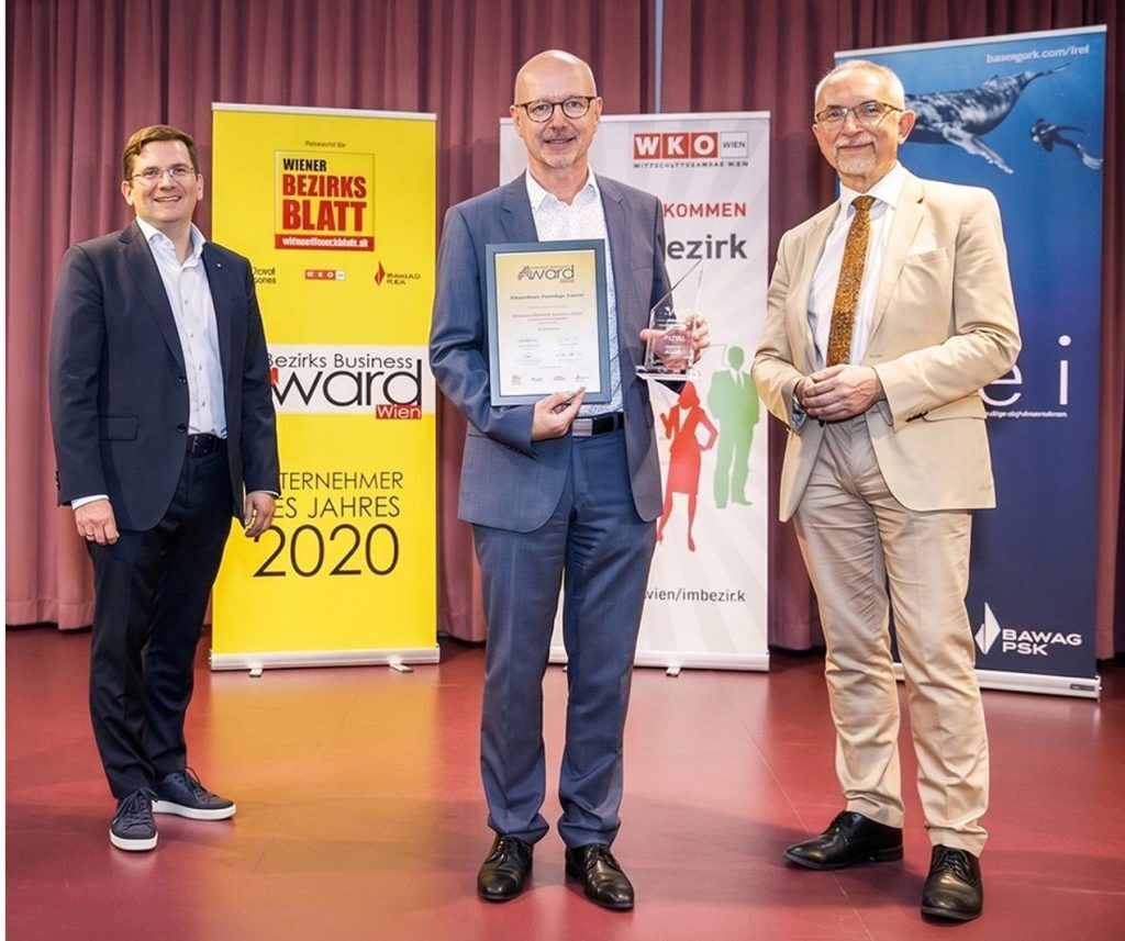 Drei Männer in Anzügen stehen auf der Bühne; der Mann in der Mitte hält eine Trophäe und eine Urkunde. Im Hintergrund sind Banner und ein Schild mit der Aufschrift "Unternehmer des Jahres 2020" zu sehen.