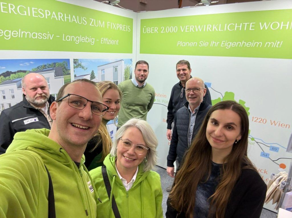 Eine Gruppe von sieben lächelnden Menschen, von denen einige grüne Kapuzenpullis tragen, posieren für ein Selfie an einem Stand der Wohnungsbauausstellung mit grünen Schildern über energieeffiziente Häuser im Hintergrund.