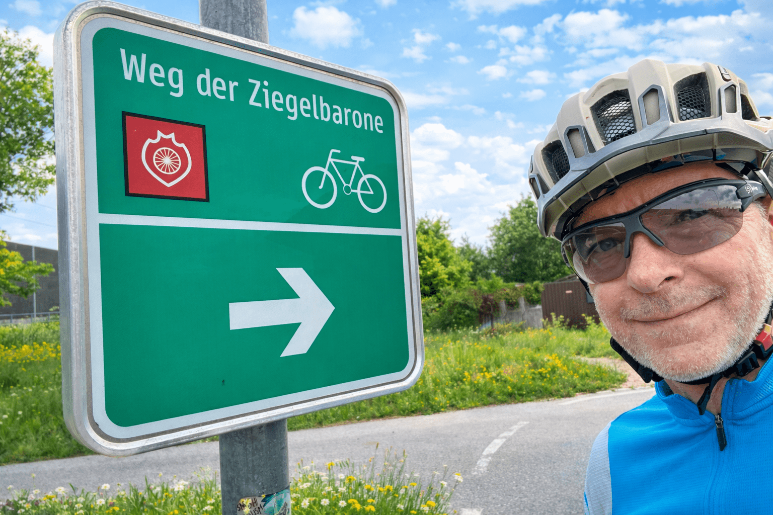 Ein Mann mit Fahrradhelm und Sonnenbrille lächelt an einem sonnigen Tag mit blauem Himmel und Wolken neben einem grünen Radwegschild mit der Aufschrift "Weg der Ziegelbarone" mit einem weißen Fahrradsymbol und einem Rechtspfeil.