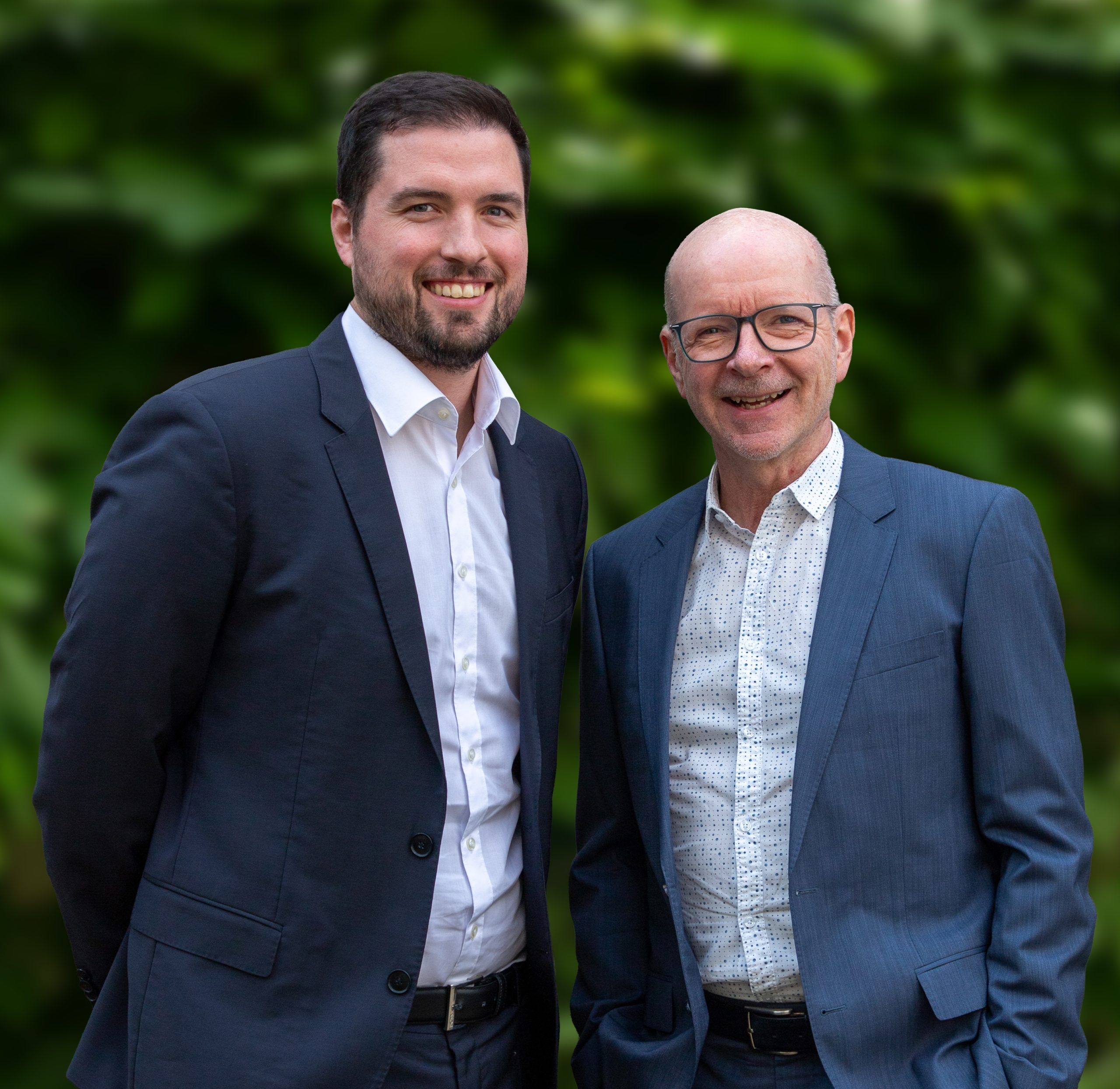 Zwei Männer in Anzügen stehen Seite an Seite und lächeln in die Kamera. Einer ist jünger, hat dunkles Haar und einen Bart, der andere ist älter, hat eine Glatze und trägt eine Brille. Der Hintergrund besteht aus unscharfem Grün.