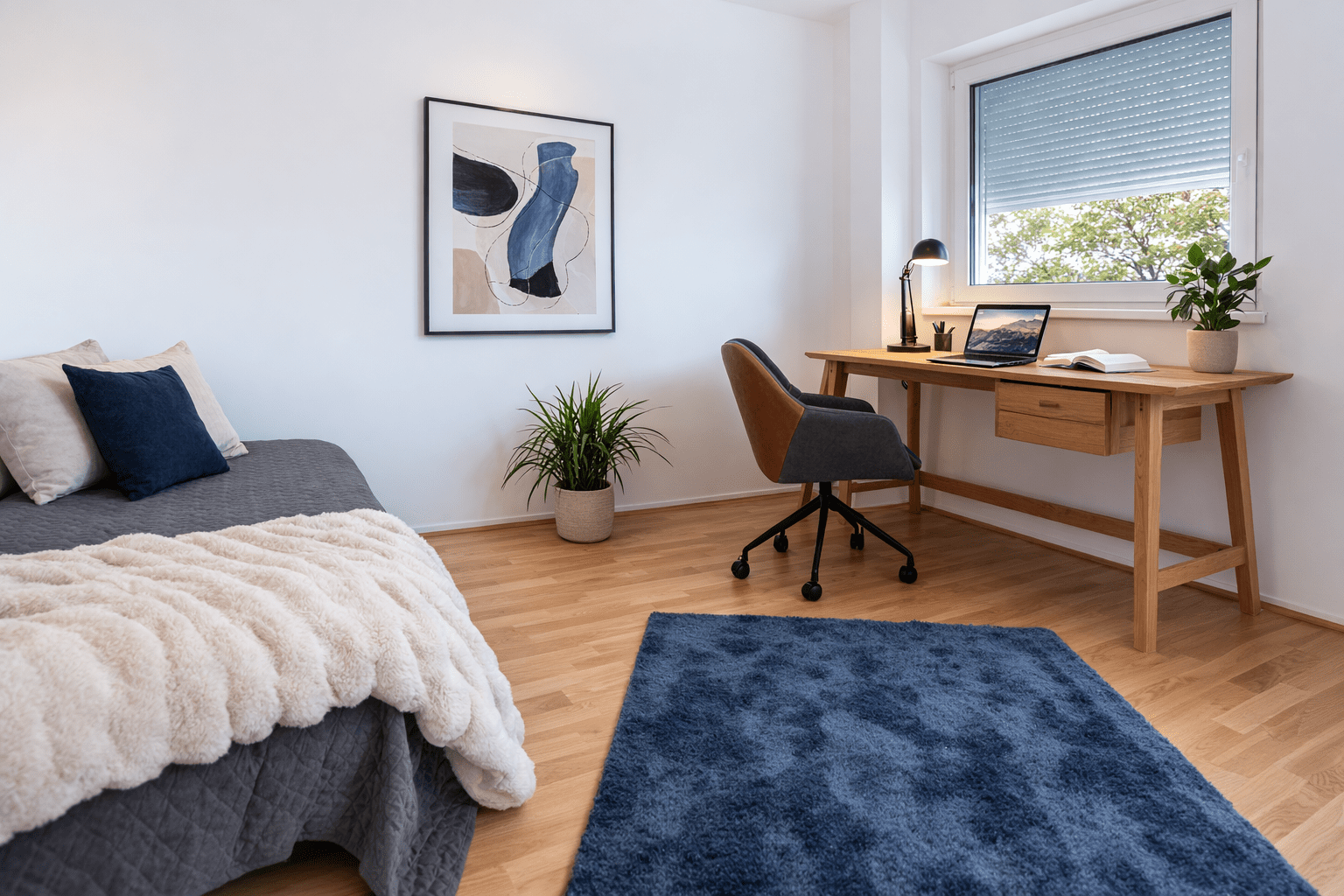 Ein modernes, aufgeräumtes Schlafzimmer mit einem Holzschreibtisch und einem Bürostuhl am Fenster, einem Laptop und einer Lampe auf dem Schreibtisch, einem Bett mit einer grauen Decke und Kissen, einer Pflanze, einem blauen Teppich und abstrakter Wandkunst.